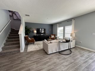 A cozy living room with a plush sofa, modern TV setup, and sleek flooring, featuring a staircase and large windows.