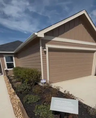 A charming single-story home in The Granary by KB Home (San Antonio, TX), featuring a brown siding exterior, a two-car garage, and a landscaped yard with an "About Our Landscaping" sign.