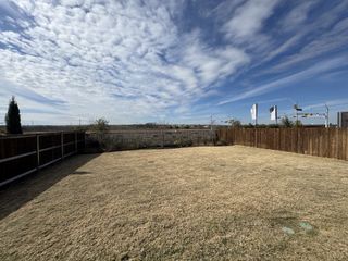 A fenced backyard with dry grass, bordered by wooden fences, under a bright sky with scattered clouds, offering a spacious outdoor area.