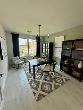 A chic home office featuring a sleek black desk, modern chandelier, geometric rug, and ample natural light through large windows.