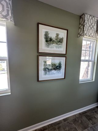 A cozy corner featuring framed landscape art, soft green walls, patterned valances, and abundant natural light through dual windows.