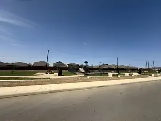 Street view A community playground in Magnolia Village (San Antonio, TX) by Rosehaven Homes features play equipment, picnic tables, and landscaping under a clear blue sky.