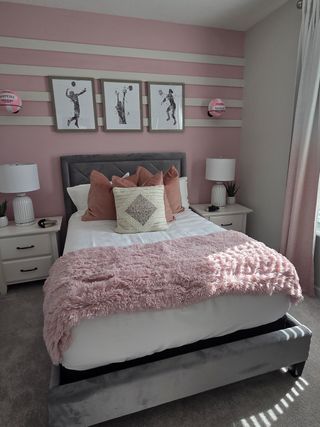 Model Home A cozy bedroom features a pink accent wall, plush bedding, framed art, and soft lighting for a welcoming ambiance.