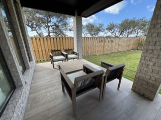 A cozy patio with modern seating, tiled flooring, and a wooden fence, overlooking a grassy yard with oak trees.