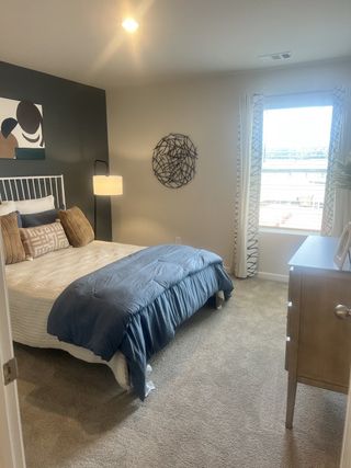 A cozy bedroom with a plush bed, modern art, stylish lamp, and natural light streaming through patterned curtains.