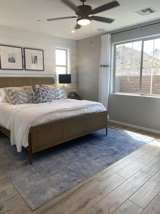 A serene bedroom features a rattan panel bed with plush bedding, a blue rug, wood-look tile flooring, framed artwork.