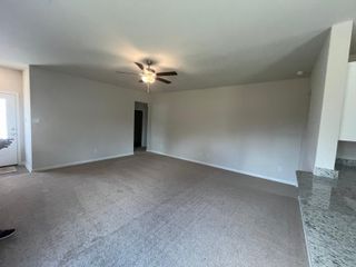 A spacious living room with plush carpet, neutral walls, and a ceiling fan, adjacent to a granite kitchen countertop.