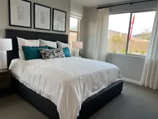 A cozy bedroom featuring a plush bed with white linens, teal accent pillows, framed artwork, and a bright window view.