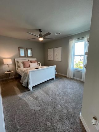 A cozy bedroom featuring a white bed, soft gray carpet, modern ceiling fan, and gentle natural light through sheer curtains.