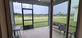 A serene screened-in patio with modern seating, overlooking lush greenery and a white picket fence in a peaceful setting.