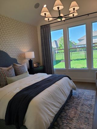 Model Home A cozy bedroom with patterned wallpaper, a plush bed, a stylish chandelier, and large windows overlooking a green backyard.