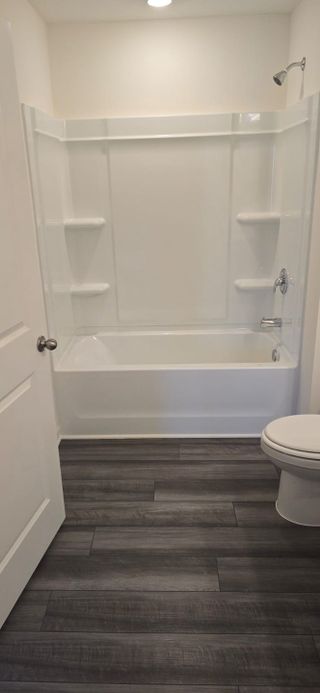 A simple white bathroom featuring a bathtub with built-in shelves, a toilet, and dark wood flooring.