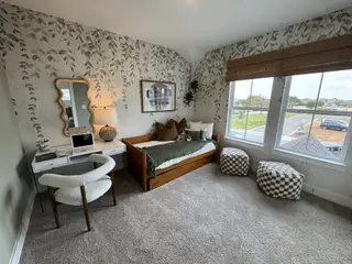 Model Home A cozy bedroom with leafy wallpaper, a wood daybed, stylish desk, and large windows with a scenic view.