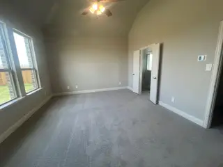 A spacious bedroom with soft carpet, neutral walls, a ceiling fan, and ample natural light from large windows.