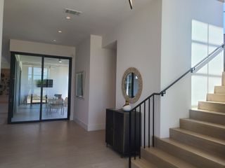 A bright entryway featuring sleek wooden stairs, a large mirror, and modern decor, leading into a glass-partitioned dining area.