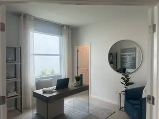 A modern home office with natural light, sleek desk, round mirror, and a cozy blue chair.
