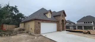 A partially completed brick and stucco home with a two-car garage. The architecture features a high-pitched roof, an arched entrance, and elegant lighting fixtures.