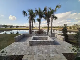A paved patio with a stone fire pit overlooks a pond and palm trees in Cross Creek by D.R. Horton (Green Cove Springs, FL), perfect for outdoor living.