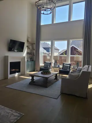 A spacious living room with a high ceiling, large windows, elegant chandelier, and cozy fireplace, blending comfort and style.