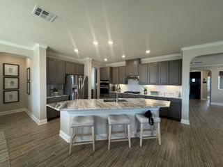 Model Home A spacious kitchen with a central island featuring a granite countertop and bar stools, gray cabinetry, stainless steel appliances, and wood-look tile flooring, opening to a hallway.