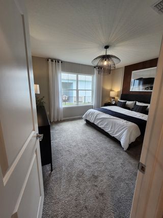 A cozy bedroom featuring soft carpeting, modern lighting, large window, and elegant bedding in a serene, minimalist style.