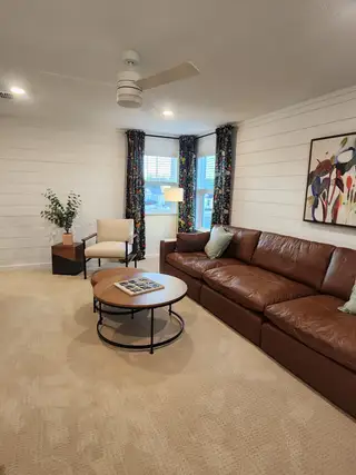 A cozy living room featuring a brown leather sofa, round coffee table, abstract art, and floral drapes.