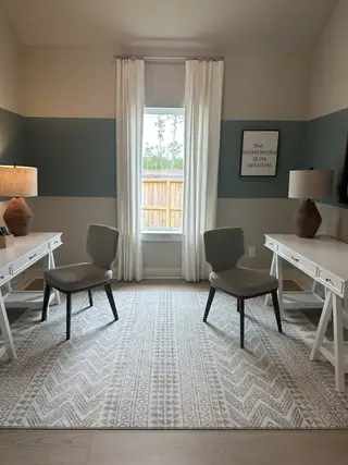 A cozy study room with dual desks, neutral decor, and ample natural light filtering through large windows.