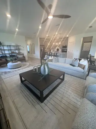 A modern living area with neutral sofas, sleek ceiling fan, stylish shelving, and open kitchen.