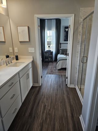 A well-appointed bathroom with dual sinks and a framed mirror opens to a bedroom with a plush bed and natural light, featuring wood-look flooring and a glass-enclosed shower.