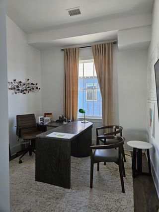 A cozy office with a wooden desk, modern chairs, and soft lighting, featuring a large window with beige curtains.