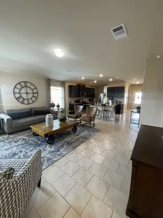Model Home A spacious living area with tile flooring, a gray sofa, a patterned rug, and a large clock flows into a kitchen with dark cabinets.