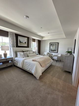 A serene bedroom with cozy carpet, a plush bed, elegant artwork, and natural light through large windows.