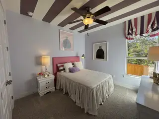 A cozy bedroom with striped ceiling, pink headboard, and modern decor, featuring a large window and elegant lighting.