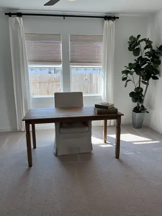 A bright home office with a wooden desk, cozy chair, and large windows draped with white curtains.