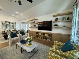 A stylish living space with a white sofa, colorful pillows, a ceiling fan, and a TV mounted on a built-in shelf wall.