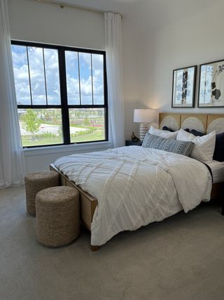 A serene bedroom featuring a cozy bed with white linens, woven stools, and abstract wall art beside a large window.
