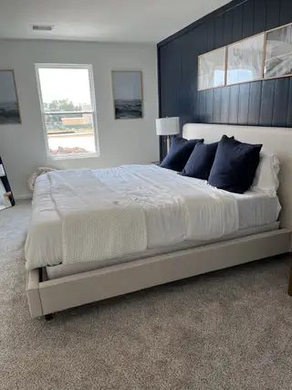 A cozy bedroom with a plush bed, dark accent wall, elegant framed artwork, and soft carpet flooring.