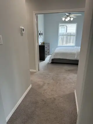 Model Home A cozy bedroom with plush carpet, soft gray walls, a modern ceiling fan, and natural light from a window.