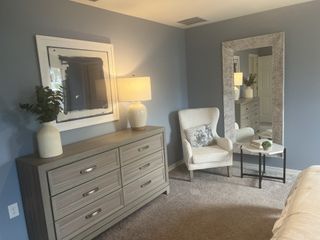 A cozy bedroom with a sleek dresser, stylish mirror, and a comfy armchair, accented by soft lighting and blue walls.