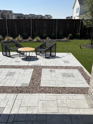 Model Home A modern patio with sleek black chairs, a wooden table, and stylish pavers overlooking a lush lawn and wooden fence.