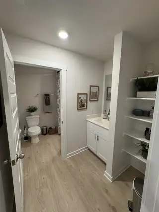 A modern bathroom with light wood flooring, white vanity, open shelving, and framed wall art for a sleek, cozy ambiance.