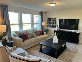 Model Home A bright and inviting living room in a Medina Crossing home, featuring a comfortable beige sofa with decorative pillows, a dark wood coffee table, a black entertainment center, and large windows (Von Ormy, TX).