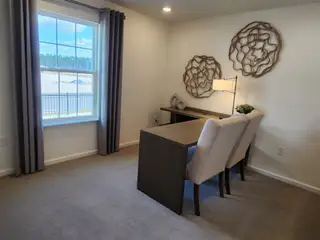 A cozy home office with soft carpeting, a wooden desk, elegant chairs, and unique wall decor. Bright window view with curtains.