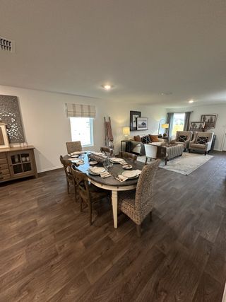 Model Home A cozy dining area with a rustic wooden table, woven chairs, and elegant decor leading into a bright, inviting living room.