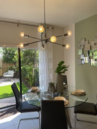 A modern dining area with a glass table, sleek chairs, and a striking chandelier, opening to a lush garden view.