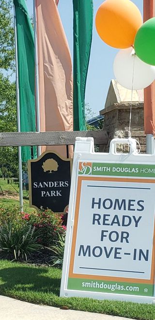 Street view A vibrant signage in Sanders Park by Smith Douglas Homes, Austell GA, promotes ready-to-move-in homes amid lush landscaping.