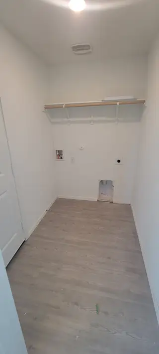 A spacious laundry room with light wood flooring, white walls, and a single shelf for storage.