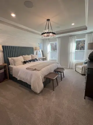 Model Home A cozy bedroom with a plush bed, elegant chandelier, and soft carpeted floor filled with natural light from large windows.