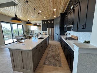 A modern kitchen with black cabinetry, wood accents, and elegant pendant lighting, featuring large windows and a spacious countertop.