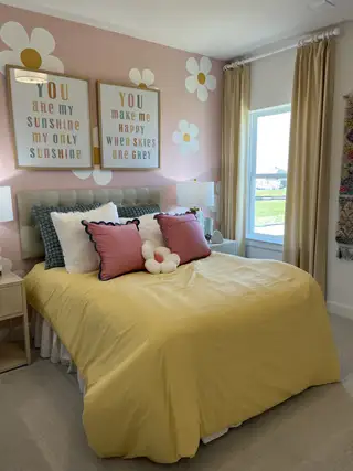 A cheerful bedroom with a yellow quilt, floral decor, soft cushions, and natural light filtering through beige curtains.